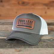 A gray and white trucker hat with a brown leather patch reading 'TRUE LEAF MARKET' on the front, displayed on a rustic wooden surface with a wood plank background.